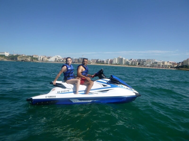 Deux personnes sur un jetski blanc et bleu naviguant sur une mer calme avec la ville en arrière-plan.