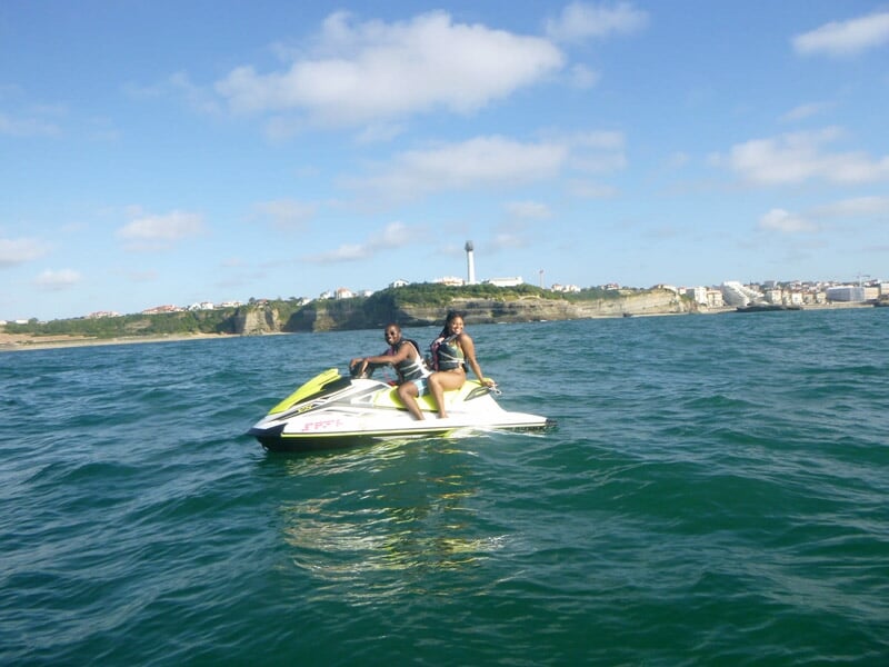 Deux personnes sur un jetski blanc et jaune naviguant sur une mer calme avec un phare visible au loin.