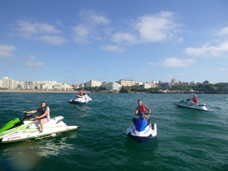 Cinq personnes sur des jetskis dispersés sur l'eau avec une ville visible en arrière-plan sous un ciel bleu.