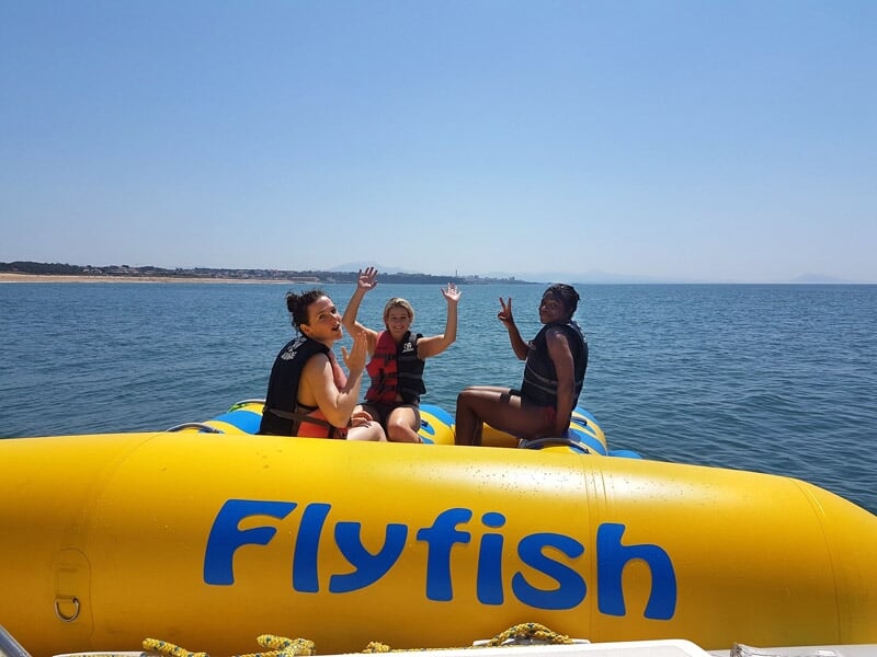 Trois personnes assises sur une grande bouée jaune nommée Flyfish, flottant sur une mer calme.