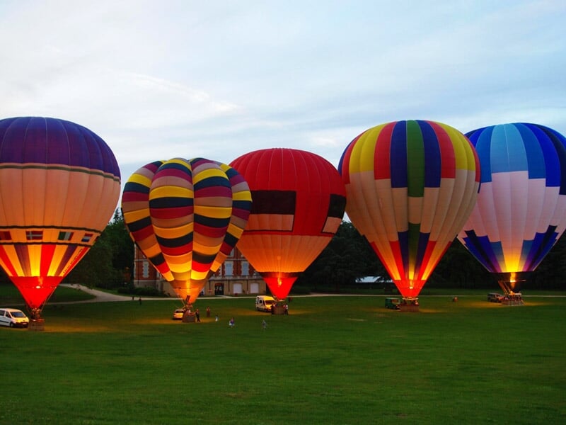 Cinq montgolfières illuminées de couleurs variées au sol lors d'un festival au crépuscule, avec un château en toile de fond