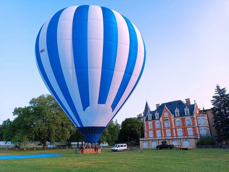 Grand ballon à air chaud aux rayures bleues et blanches survolant un château avec un ciel clair en arrière-plan