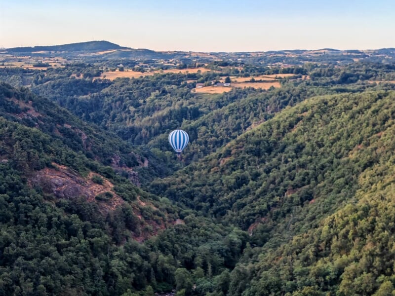 Montgolfière bleue et blanche survolant une vallée boisée avec collines et champs sous un ciel dégagé.