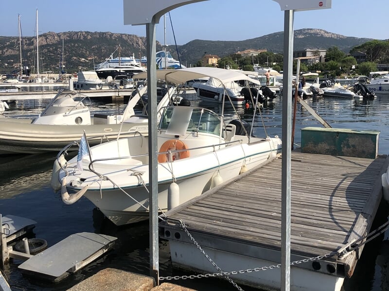 Bateau école blanc amarré à un ponton en bois, avec bouée orange et drapeau bleu, vue sur le port et collines.
