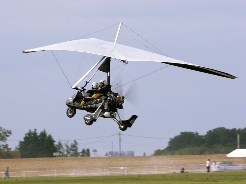 ULM à aile delta blanc en vol au-dessus d'un champ avec un pilote et un passager portant un casque.