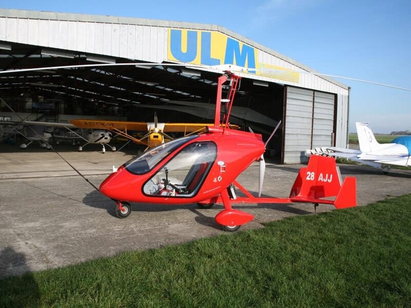Gyroptère rouge garé devant un hangar marqué ULM, avec un ciel clair en arrière-plan.