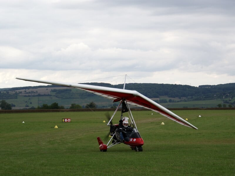 ULM à aile delta rouge avec un pilote en casque volant au-dessus d'un champ vert et collines au loin.