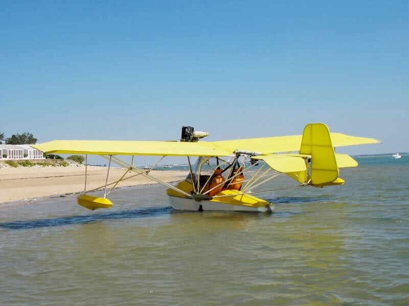 Hydravion jaune amarré près d'une plage de sable sous un ciel bleu clair. L'avion est posé sur l'eau peu profonde, ses flotteurs touchant à peine la surface. En arrière-plan, une maison blanche entour