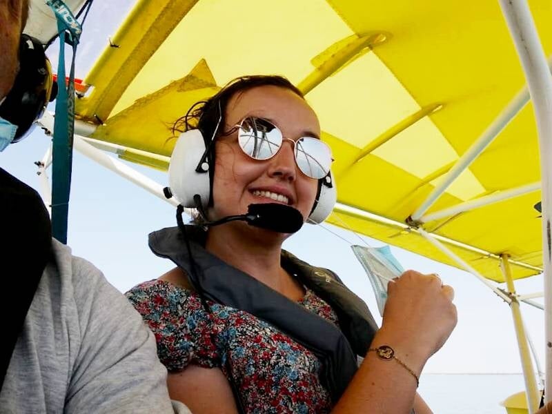 Femme souriante portant des lunettes de soleil réfléchissantes et un casque de communication avec micro, assise dans un avion léger. Elle est vêtue d'une blouse à motifs floraux et porte un gilet de s