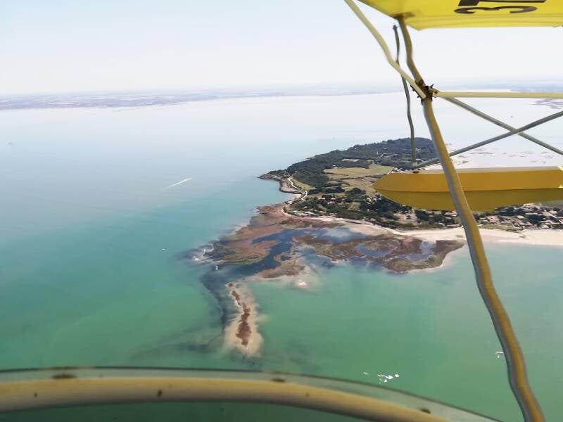 Vue aérienne d'une côte bordée par une mer turquoise avec des nuances vert émeraude. Une étendue de terre verte et brune, composée de prairies et de zones marécageuses, s'avance dans l'eau. L'aileron 