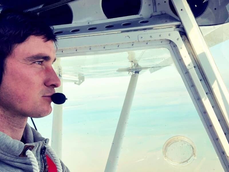 Homme concentré portant un casque avec micro, aux commandes d'un avion ultraléger en vol au-dessus de la côte.