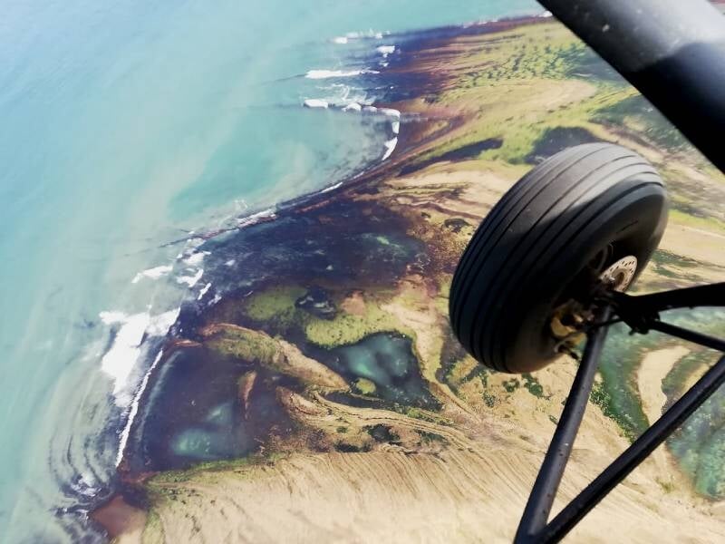Vue aérienne d'une côte rocheuse et verdoyante prise depuis un avion ultraléger en vol, avec une roue visible.