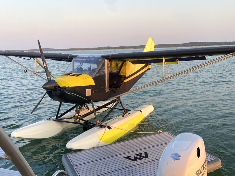 Petit avion ULM jaune et noir équipé de flotteurs, amarré sur l'eau calme d'un port au coucher du soleil.