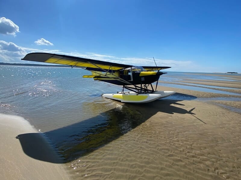 Avion ULM jaune et noir posé sur des flotteurs sur une plage de sable avec ciel bleu et mer calme en arrière-plan.