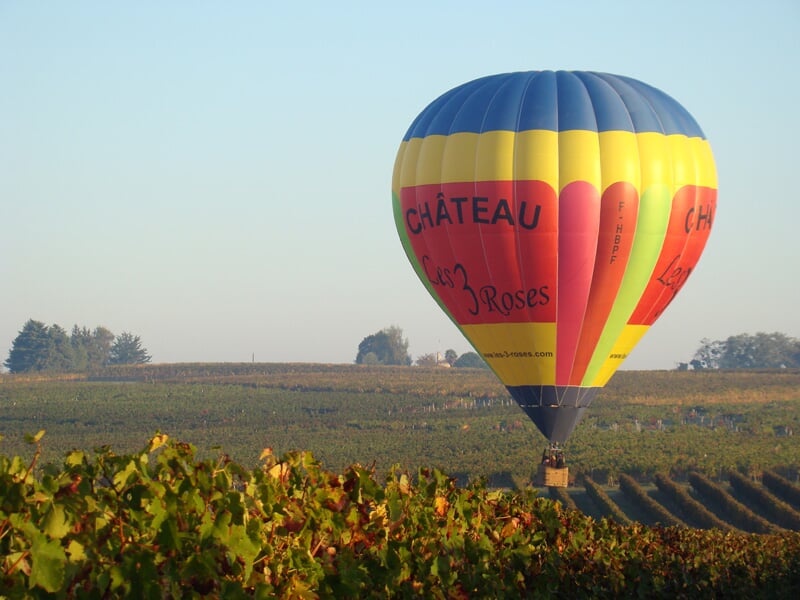 Montgolfière colorée jaune, rouge et bleu survolant un vignoble au lever du jour dans un paysage rural paisible.