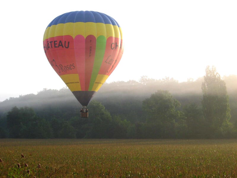 Montgolfière colorée volant au-dessus d'un champ avec une légère brume matinale et des arbres en arrière-plan.