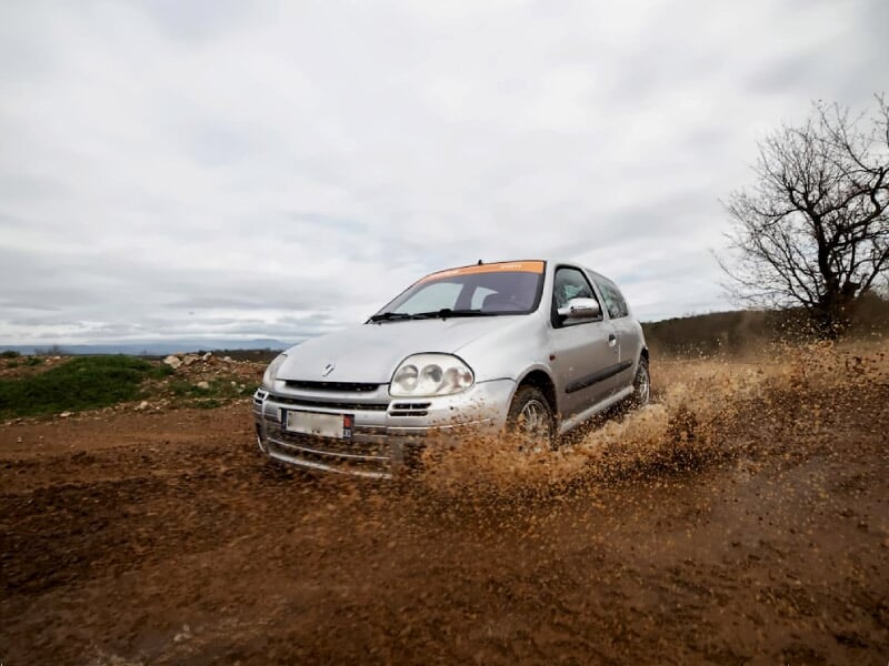 Renault grise roulant sur un chemin boueux, éclaboussant l’eau, sous un ciel nuageux avec un arbre sans feuilles à droite.