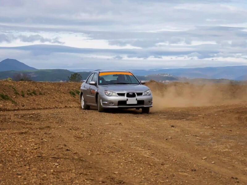 Voiture grise avec bandeau orange sur le pare-brise roulant sur un chemin de terre en lacet, soulevant un nuage de poussière, avec collines et ciel nuageux en arrière-plan.