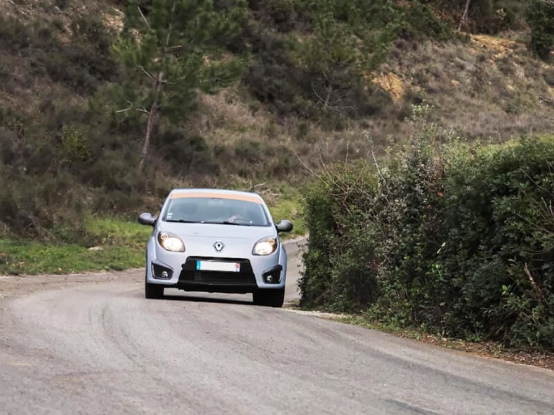 Renault grise prenant un virage sur une route étroite bordée de buissons, avec un conducteur visible à l’intérieur.