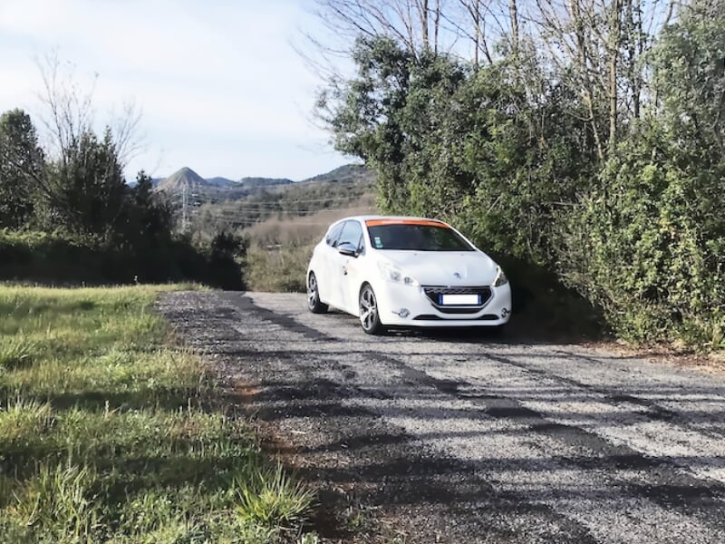Peugeot blanche en approche sur une route de campagne asphaltée, entourée de verdure et collines au loin.