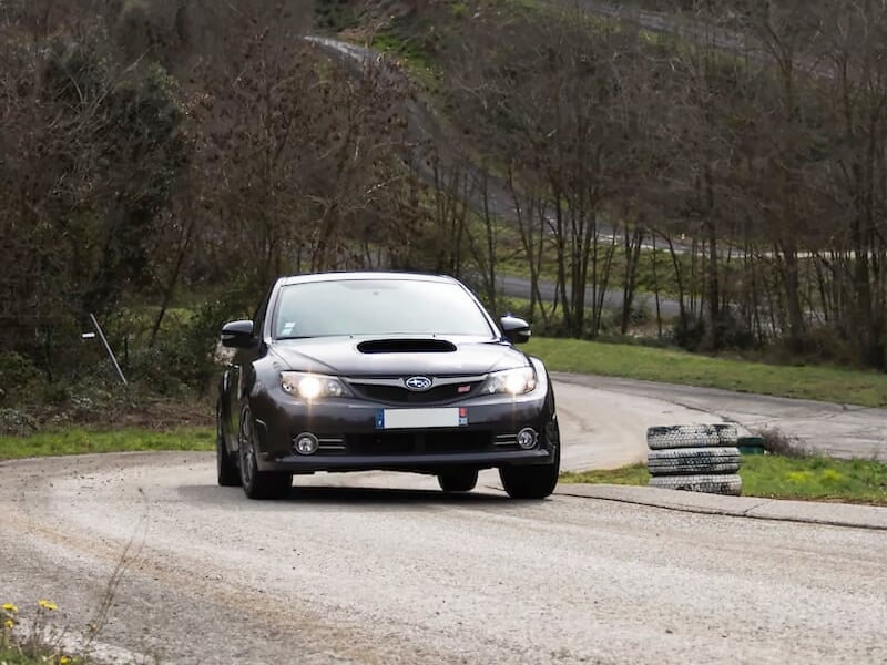 Subaru noire en virage sur une route sinueuse bordée d’arbres dénudés, avec des pneus empilés sur le bas-côté.
