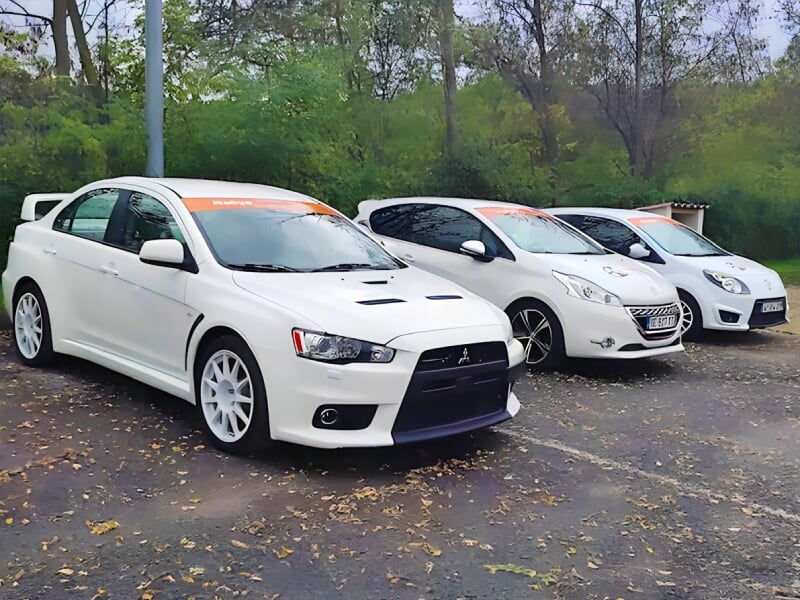 Quatre voitures blanches garées côte à côte sur un parking boisé, dont une Mitsubishi sportive au premier plan, toutes avec bandeau orange sur le pare-brise.