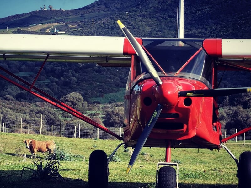 Avion rouge au sol dans un champ vert avec des collines en arrière-plan sous un ciel bleu clair