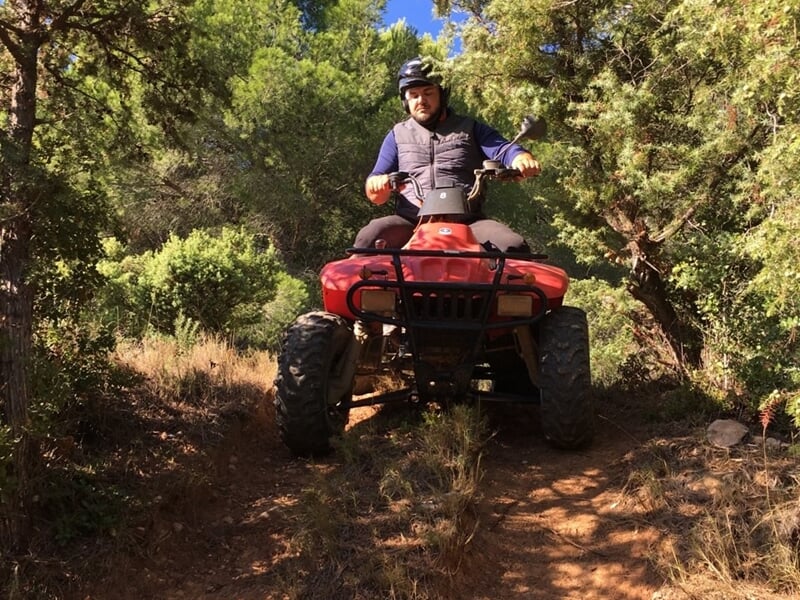 Homme en tenue de protection conduisant un quad rouge sur un chemin étroit en forêt dense sous le soleil.