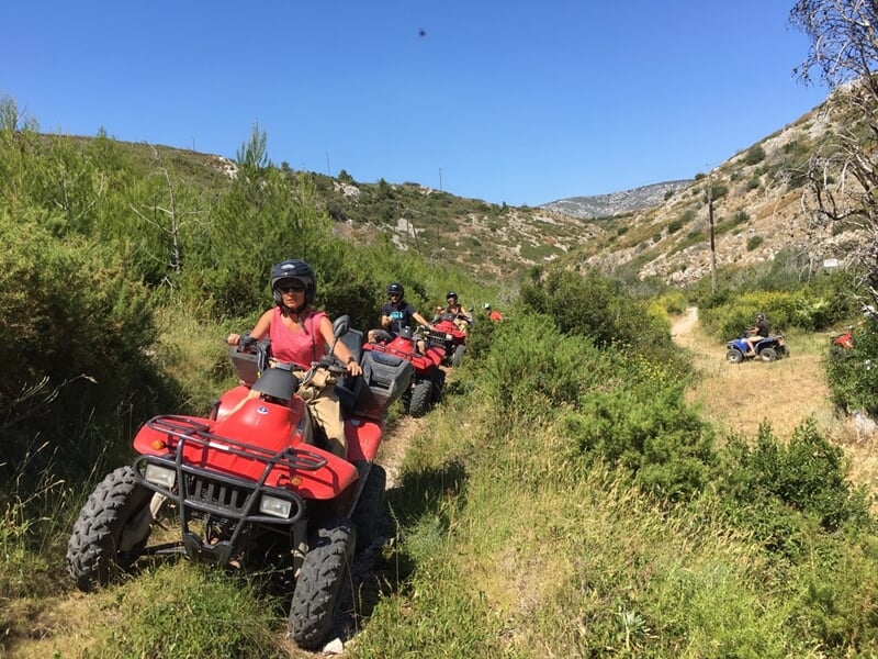 Groupe de personnes en quad rouge roulant en file indienne sur un sentier entouré de végétation et collines.
