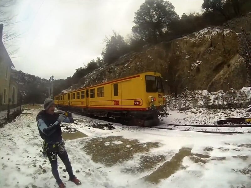 Personne en combinaison de canyoning debout sur un sol enneigé près d'un train jaune dans un paysage montagneux.