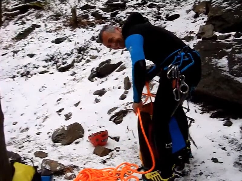 Homme en tenue de canyoning avec casque rouge et matériel préparant une corde dans un environnement enneigé.