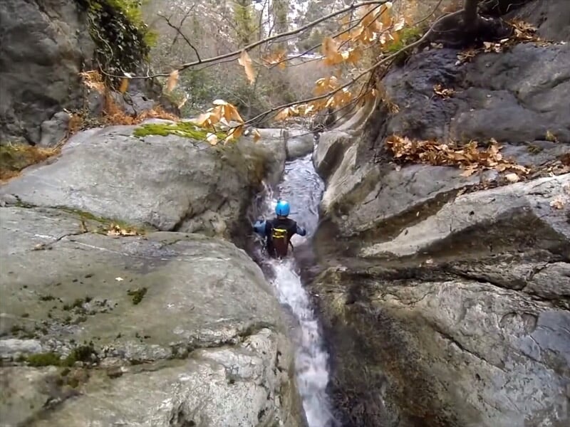 Personne en combinaison et casque rouge marchant dans un canyon étroit avec des rochers et de la végétation.