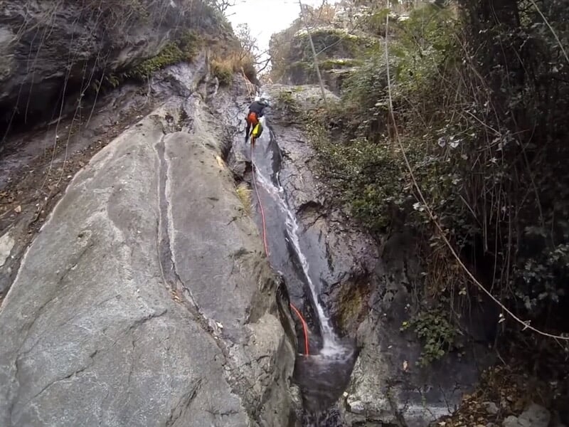 Personne en combinaison bleue descendant en rappel une cascade d'eau dans un canyon rocheux étroit et escarpé.