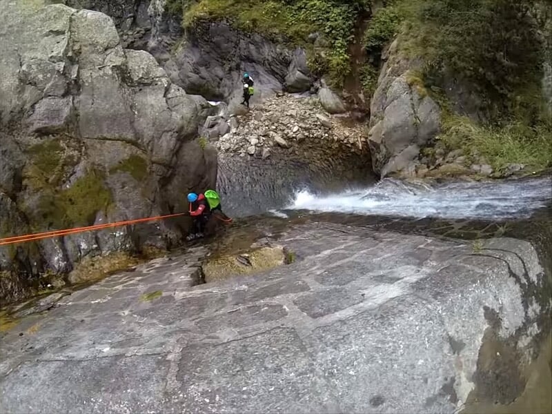Personnes en combinaisons et casques rouges marchant dans un canyon rocheux étroit avec de l'eau et des rochers.