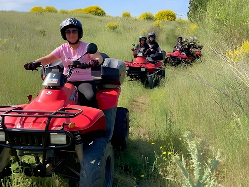 Personne en casque et lunettes conduisant un quad rouge sur un chemin de terre entouré de végétation verte et fleurs.