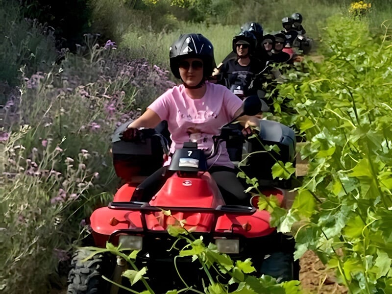 Personne en casque noir conduisant un quad rouge sur un chemin étroit bordé de fleurs et de végétation dense.