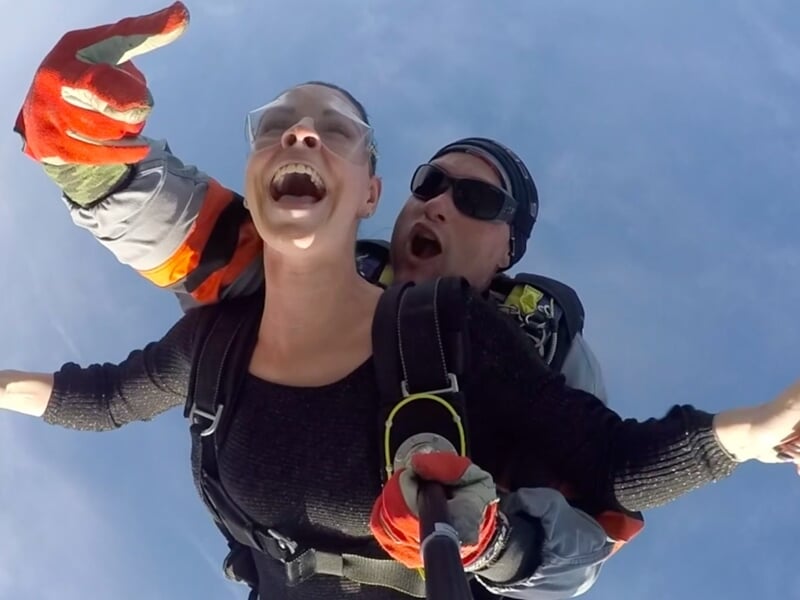 Saut en parachute tandem, deux personnes souriantes en chute libre au-dessus de la campagne.