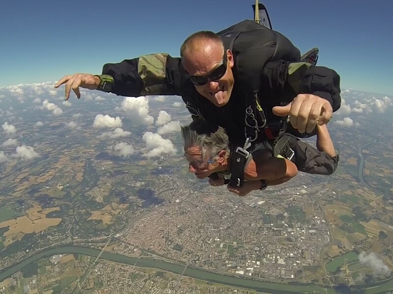 Parachutiste en tandem souriant en chute libre, paysage vert et ciel bleu en arrière-plan.