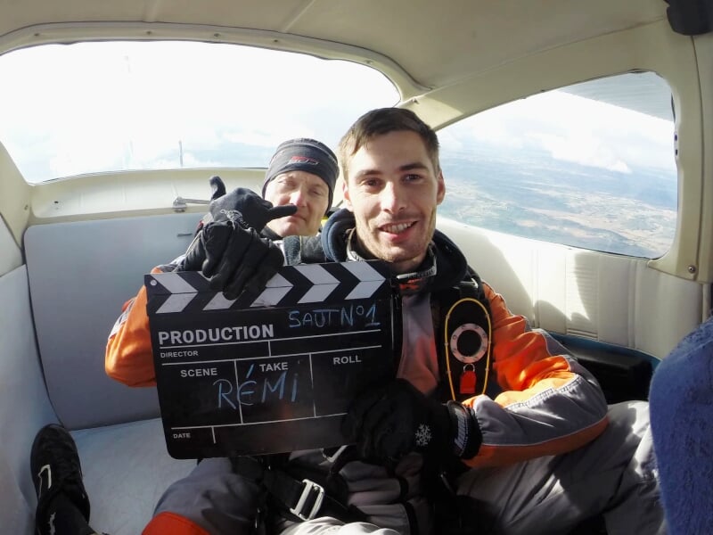 Deux hommes en combinaison de saut en parachute dans un avion, tenant une claquette de cinéma avec le nom Rémi.