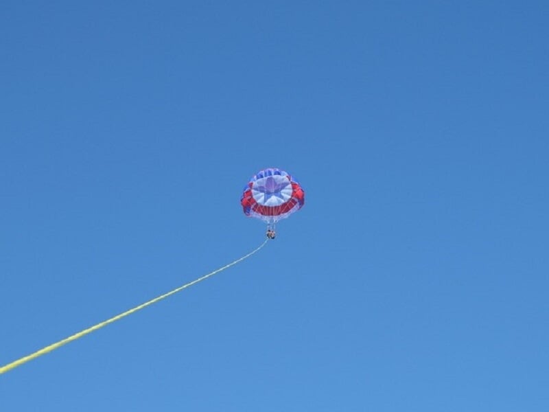 Parachute ascensionnel rouge, blanc et bleu avec trois personnes suspendues, vu de loin dans le ciel.