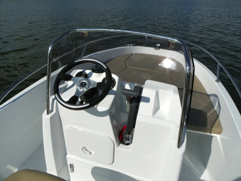 Intérieur d'un bateau blanc avec volant, levier de vitesse et sièges beiges, sur une eau calme.