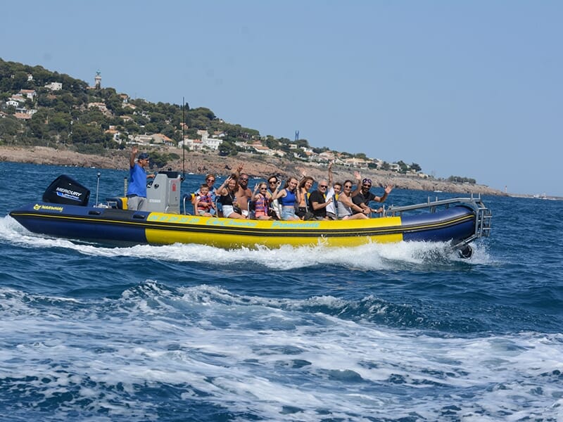 Un groupe de vacanciers dans un bateau bleu et jaune naviguent en mer avec la côte rocheuse en arrière-plan