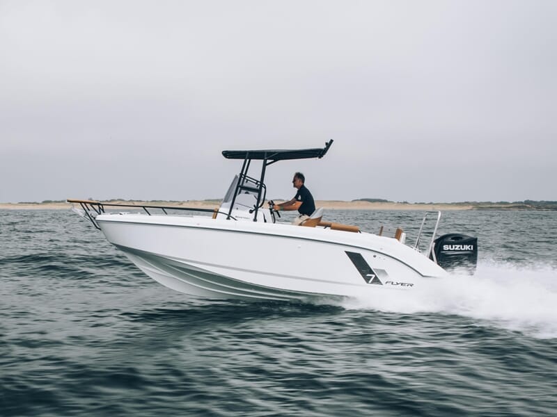 Un homme seul conduit un bateau blanc Beneteau Flyer 7 à moteur Suzuki, équipé d’un toit rigide, filant sur une mer grise proche d’un rivage sablonneux.