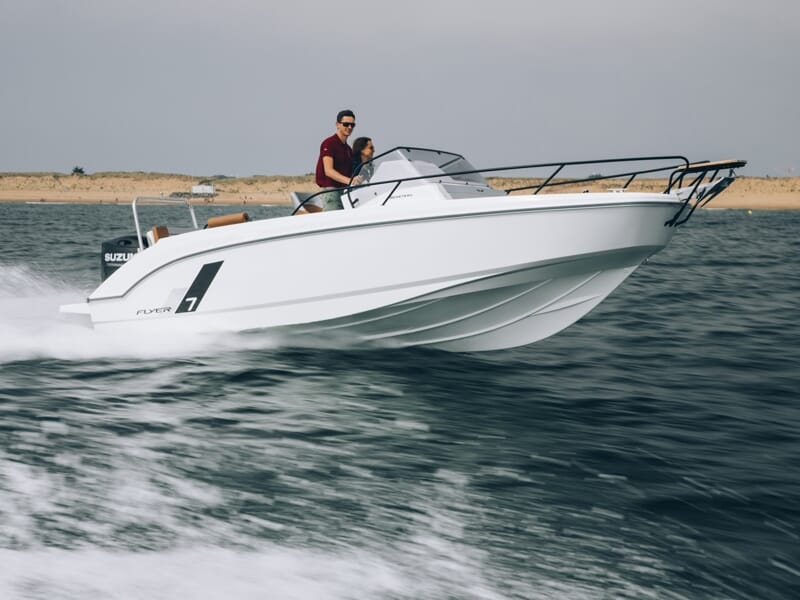 Un couple navigue sur un bateau blanc Beneteau Flyer 7 à moteur Suzuki, avançant rapidement sur une mer légèrement agitée près d’une plage sablonneuse.