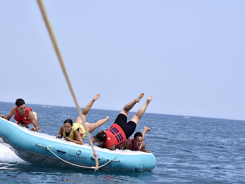 Quatre personnes portant des gilets de sauvetage rouges et jaunes sur une bouée tractée bleue en mer, s'amusant sous un ciel clair.