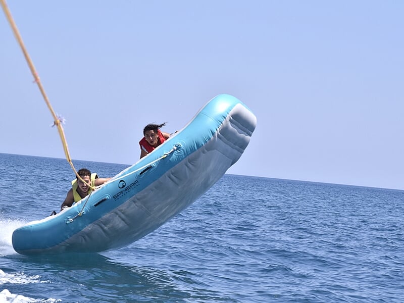 Bouée tractée bleue en mer avec quatre personnes en gilets de sauvetage, la bouée est en train de se soulever hors de l'eau sous un ciel dégagé.