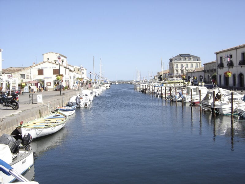 Vue d'un port avec bateaux amarrés de chaque côté d'un canal, bâtiments blancs et ciel bleu clair.
