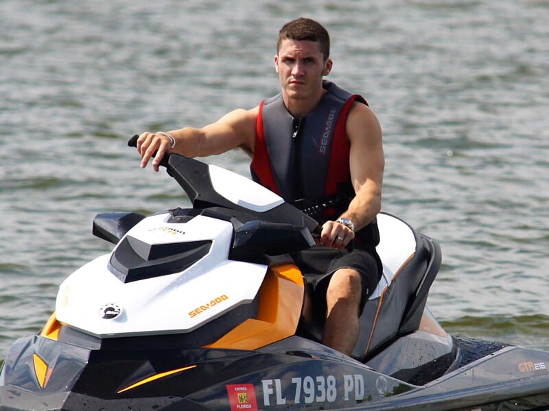 Homme en gilet de sauvetage noir conduisant un jetski blanc et orange sur une eau calme.