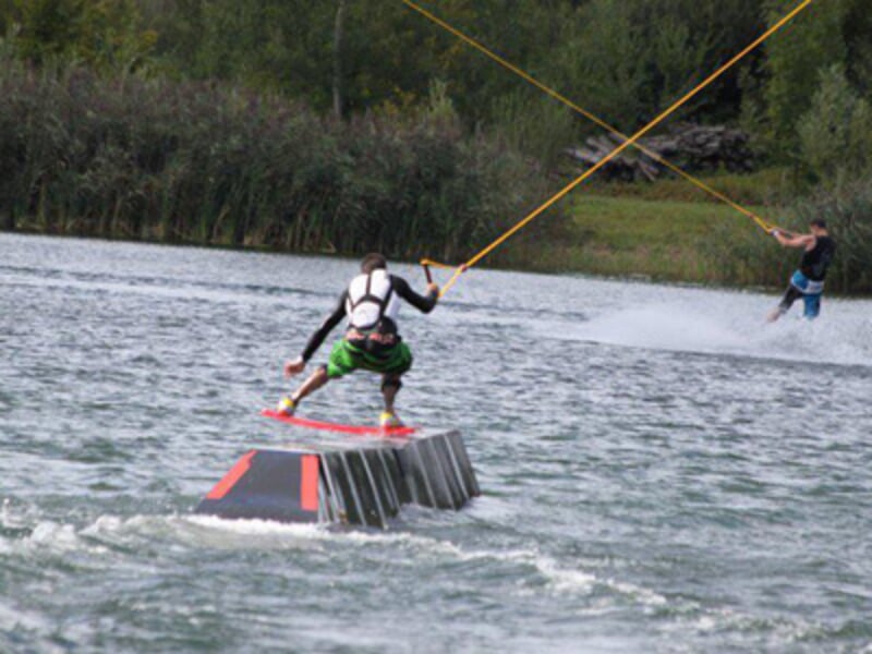 Un wakeboarder effectue un saut sur un obstacle métallique dans un lac entouré d'arbres verts en arrière-plan.