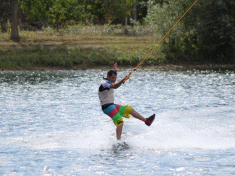 Un homme en short coloré pratique le wakeboard en glissant sur l'eau, tenant la poignée du câble avec une main levée.